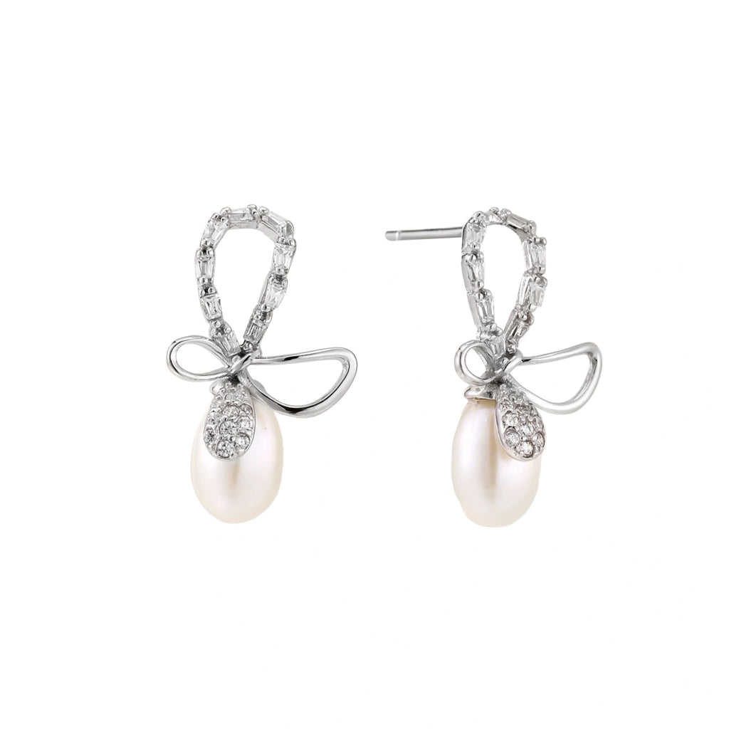 Silver bow-shaped stud earrings with baguette crystals and dangling white freshwater pearls on cream background