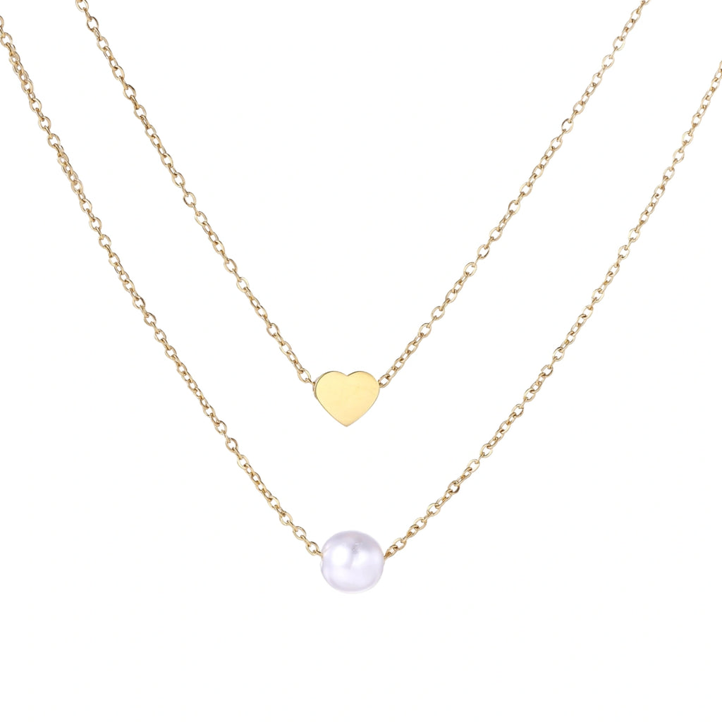Two delicate gold-plated chains with heart charm and white pearl bead on cream background, minimal layered design.