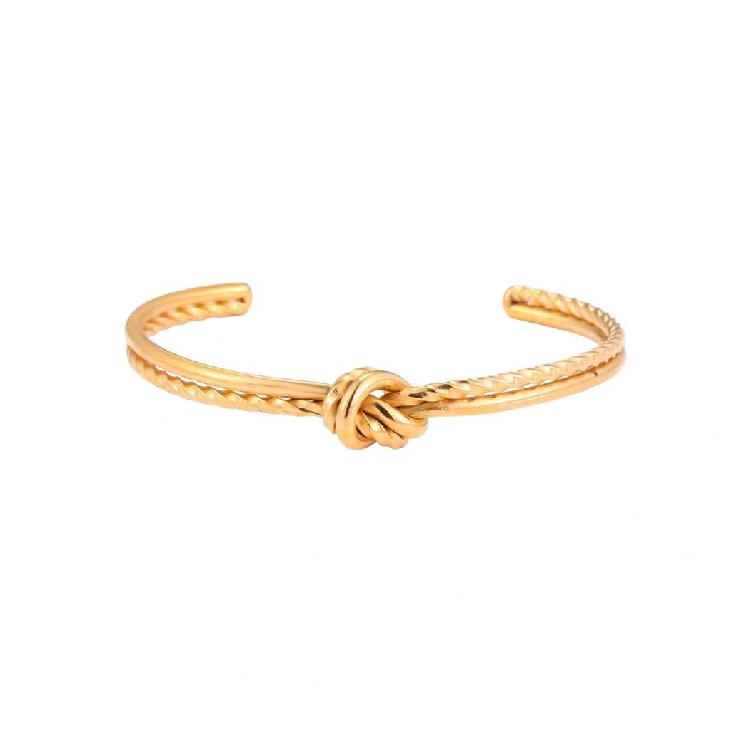 Gold cuff bracelet with twisted rope texture and decorative knot centerpiece on cream background.