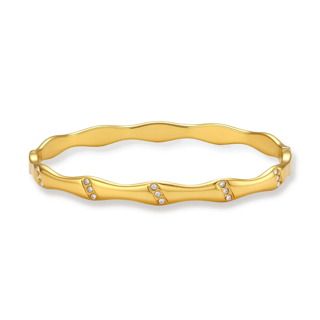 Gold-toned wavy bangle with bamboo-style segments and CZ crystal clusters on cream background. 18K plated stainless steel.